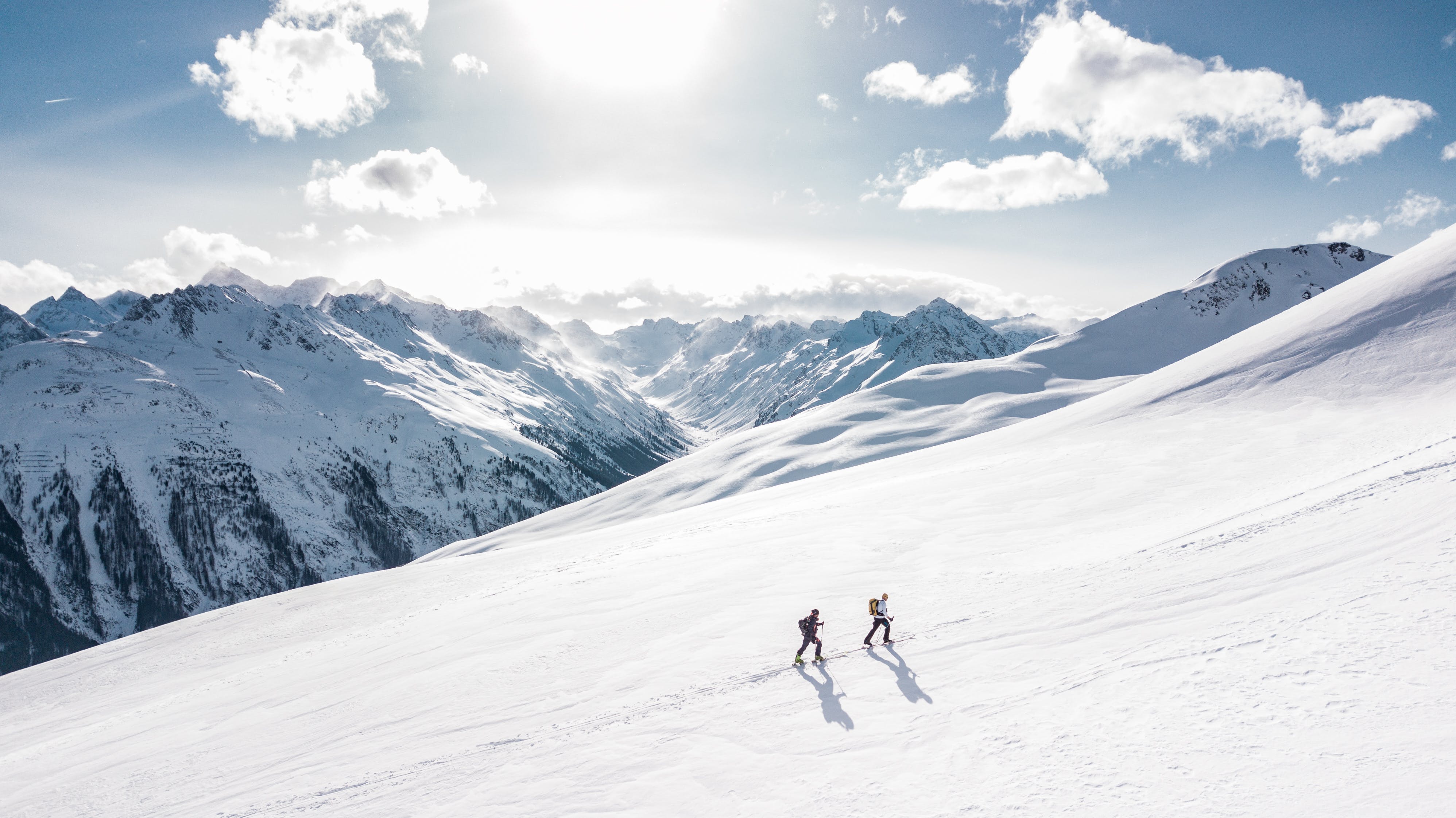 Two mountain Climbers traipsing up a snow covered hill in the mountains, with bright sunlight illustrating views across multiple mountain ranges, bright blue sky punctuated by small clouds.
