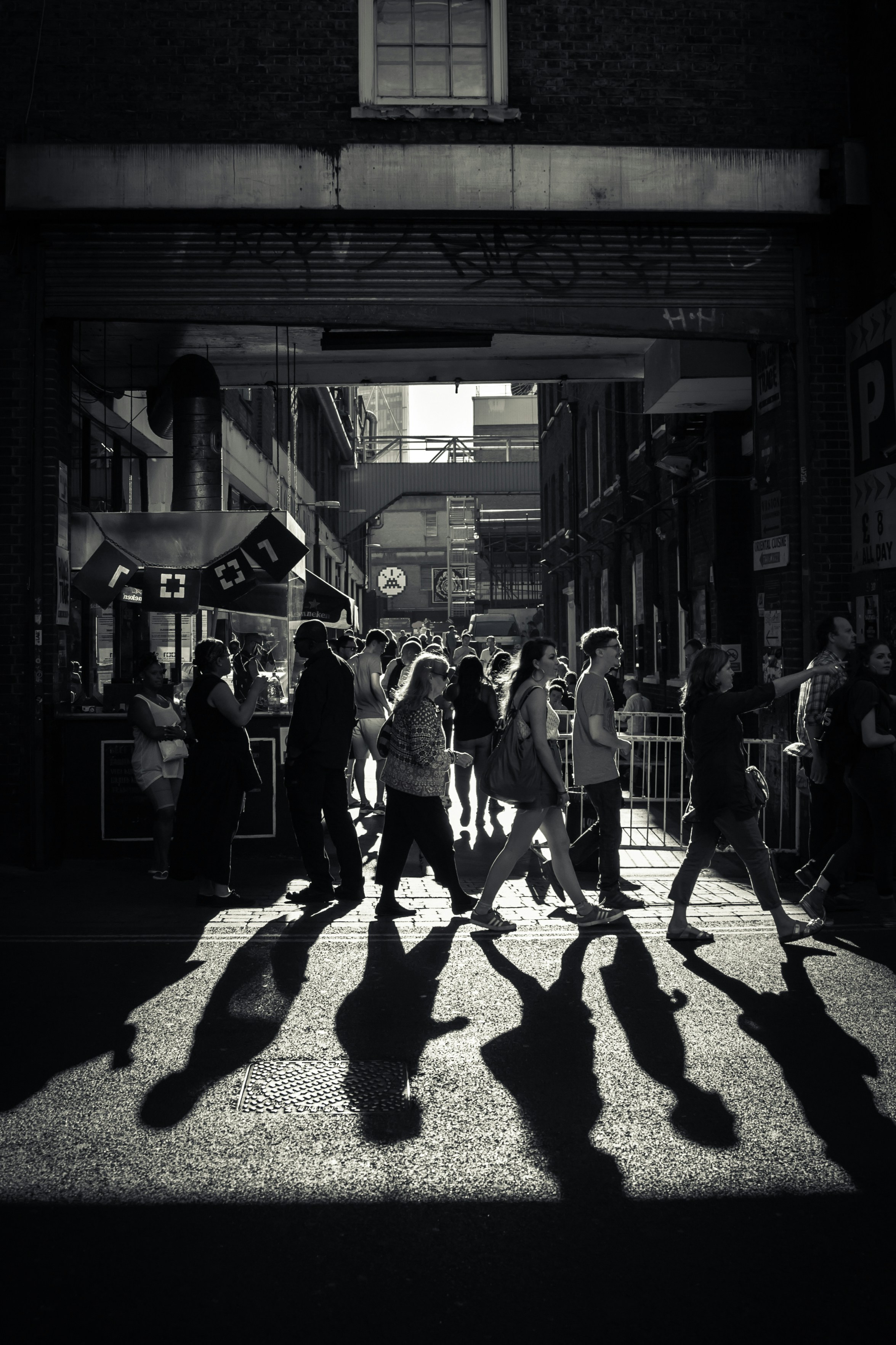 Black & white photo of random people walking in a crowded city street, with sunlight behind them casting long shadows. A sense of movement, activity and a sense of summer, due to the people walking shorts, skirts and t-shirts.